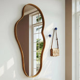 Elegant wavy mirror on a white wall with a bench and handbag in the foreground.