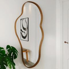 Wooden floor mirror with wavy design on a white wall with abstract art and plant.