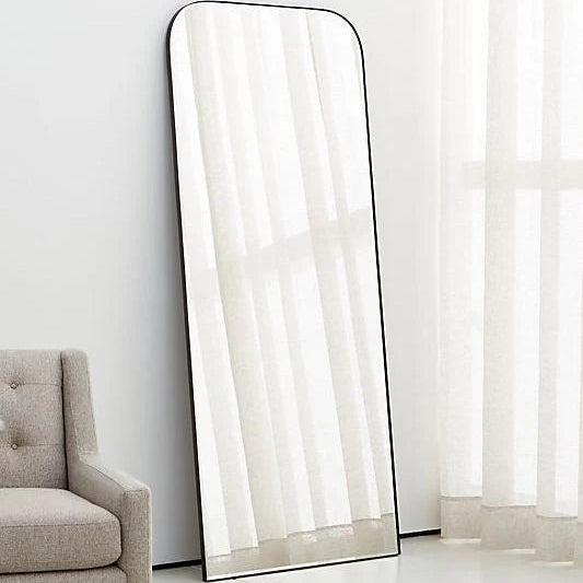 Tall floor mirror with black frame leaning against a white wall next to a gray armchair.