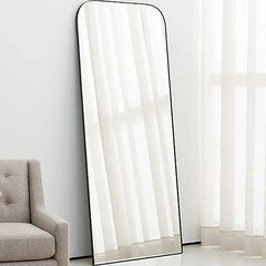Tall floor mirror with black frame leaning against a white wall next to a gray armchair.