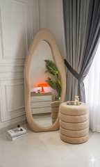 Beige floor mirror with a cylindrical stool in a room with white walls and gray curtains.