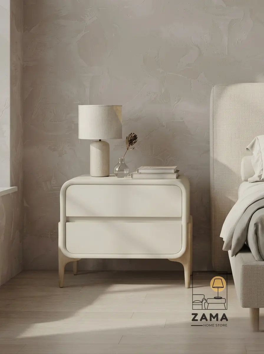 Contemporary two-drawer curved nightstand in cream finish, styled with a lamp and books on the left side of a neutral bedroom next to a bed