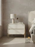 Contemporary two-drawer curved nightstand in cream finish, styled with a lamp and books on the left side of a neutral bedroom next to a bed