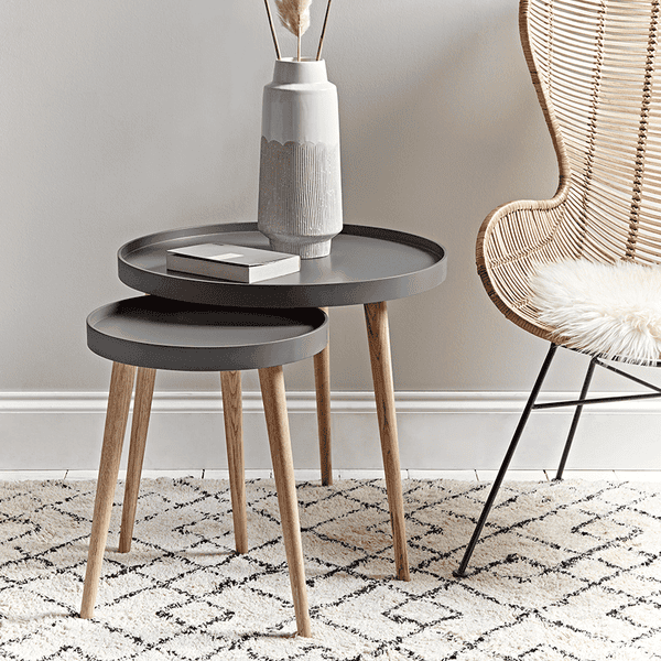 Nest of two dark gray side tables with wooden legs, a vase with dried plants, and a wicker chair in a room.