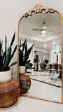 Large ornate mirror reflecting a modern kitchen with a wicker stool and potted plant.